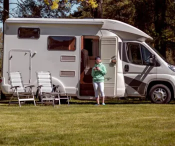 Frau steht mit Kaffeetasse vor ihrem Camperwagen umgeben von Bäumen