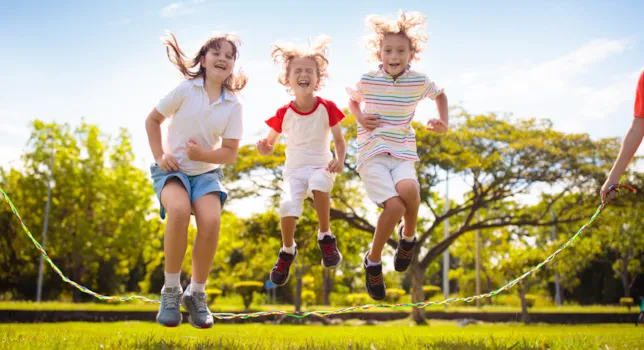 Drei Kinder im Grundschulalter springen zusammen Seil auf einer Wiese