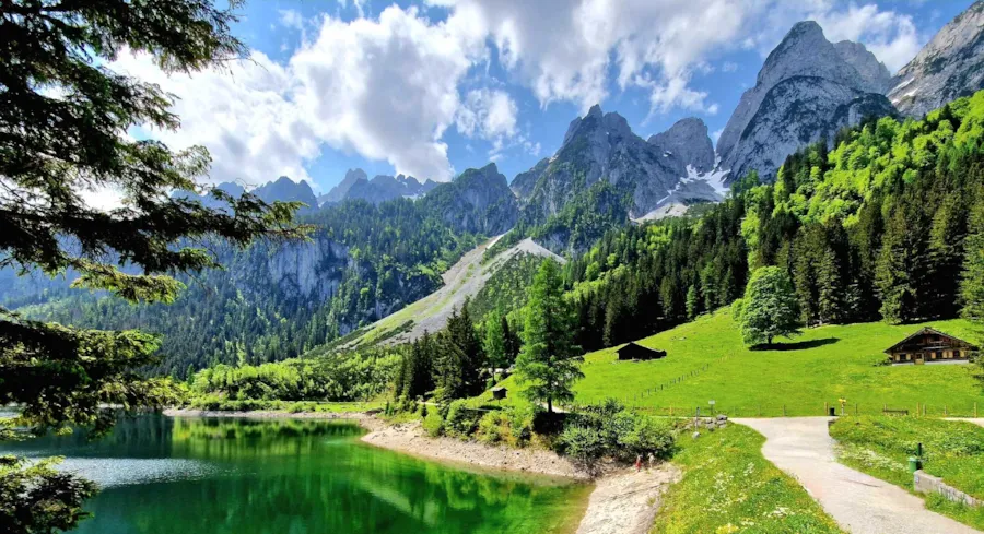 Een berglandschap met een turquoise kleurig meer aan de linkerzijde en een wandel- en fietspad eromheen. Wolken in de lucht en naaldbomen in de verte | Camping Direct 