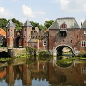 Een oude stadspoort met torens en vestingmuren, gebouwd van baksteen. De poort heeft grote bogen waar water onderdoor loopt | Camping Direct