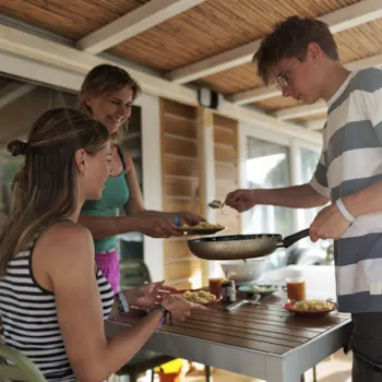 Moeder met een tienerzoon en -dochter aan tafel op het terras van een stacaravan. De jongen schept het eten op.