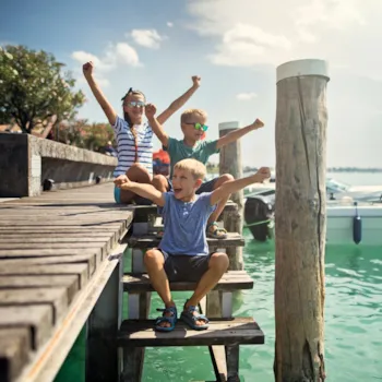 Drie kinderen zitten met de armen in de lucht op de trap van een houten steiger, met op de achtergrond boten.