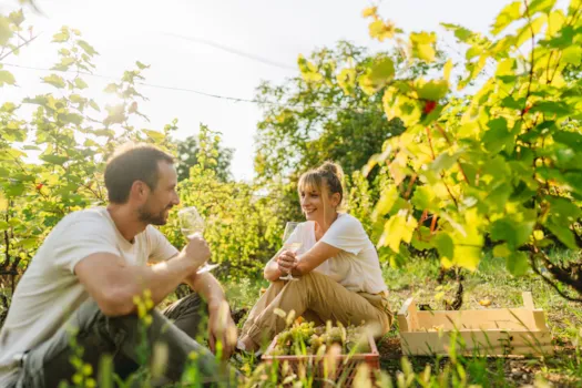 Paar entspannt im Weinberg, hält Wein-Gläser und lächelt sich an.