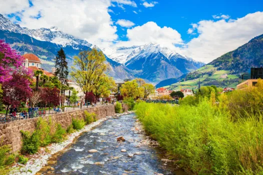 Bergstadt Meran in Südtirol mit Flusslauf, Promenade, blühenden Bäumen und schneebedeckten Alpen im Hintergrund unter blauem Himmel.