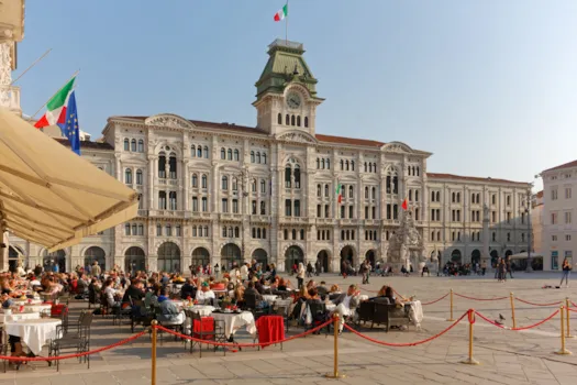 Gefüllter AUßenbereich des Caffè degli Specchi in Triest, im Hintergrund das Rathaus der Stadt 