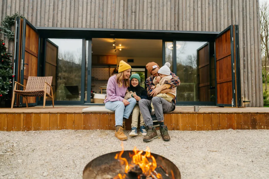 Familie mit zwei kleinen Kindern im Herbst vor einem Mobilheim auf der Terrasse sitzend und gemeinsam lachend, im Vordergrund brennt eine Feuerschale