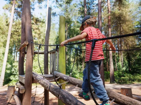 Ein Kind balanciert vorsichtig auf einer Hängeholzbrücke umgeben von hohen Bäumen in einem sonnigen Waldpark