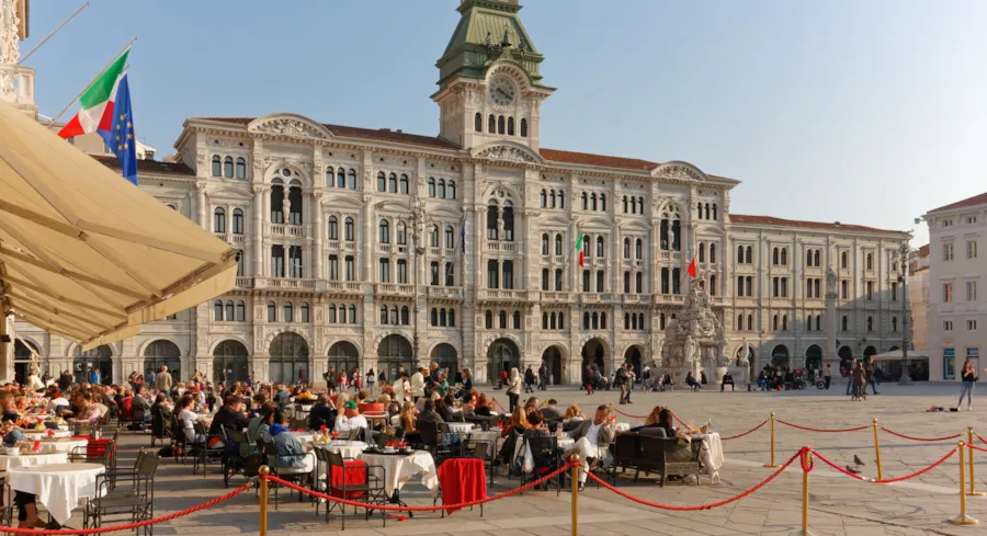 Gefüllter Außenbereich des Caffè degli Specchi in Triest, im Hintergrund das Rathaus der Stadt 