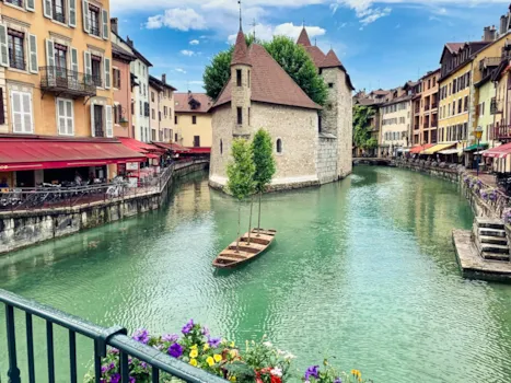 Malersicher Kanal in Annecy mit türkisfarbenem Wasser, Holzbooten und einem historischen Steingebäude in der Mitte, gesäumt von farbenfrohen Häusern und Cafés.