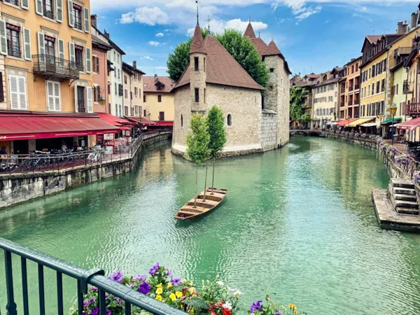 Malersicher Kanal in Annecy mit türkisfarbenem Wasser, Holzbooten und einem historischen Steingebäude in der Mitte, gesäumt von farbenfrohen Häusern und Cafés.
