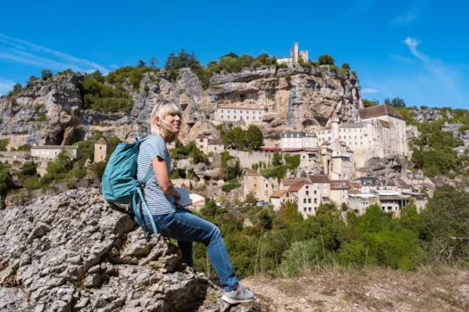 Seniorin mit Rucksack sitzt auf einem Felsen und blickt auf das in eine Felswand gebaute Dorf Rocamadour in der Dordogne, Frankreich.