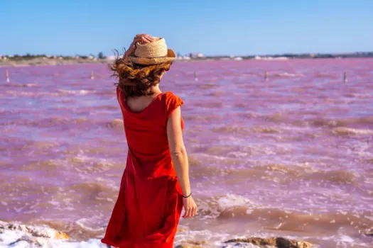 Frau in rotem Kleid und mit Hut steht im Wind an der rosa Lagune von Torrevieja, Alicante. Spanien, Urlaub am Mittelmeer.