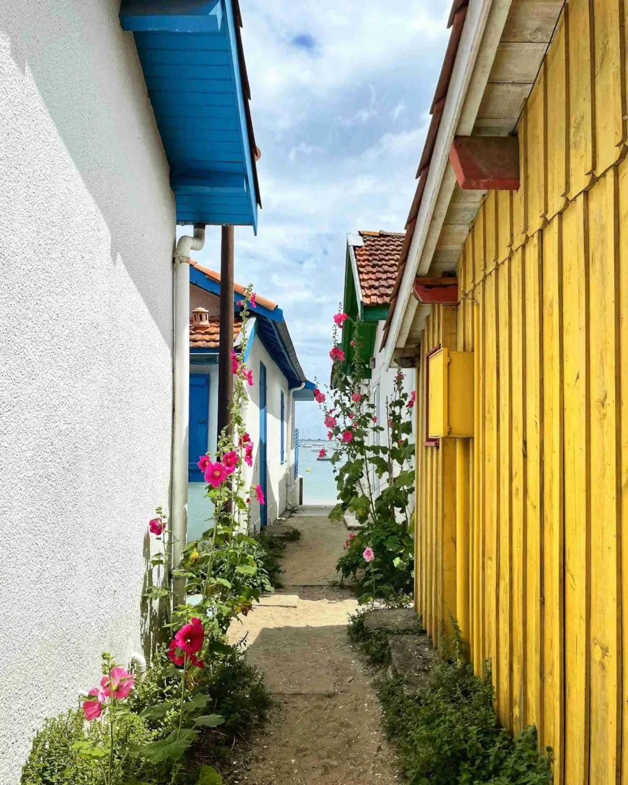 Blick durch kleine, von bunten Häusern gesäumte, Gasse auf das Bassin in Cap Ferret