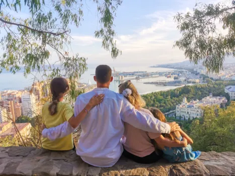Rückansicht einer Familie, die gemeinsam auf einem Aussichtspunkt auf einem Hügel sitzt und den Panoramablick auf Málaga genießt