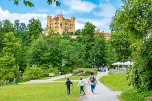 Blick auf einen Park mit Schloss Hohenschwangau im Hintergrund, mit Radfahrern und Fußgängern, Gastronomie für Touristen, Allgäu, Bayern, Deutschland