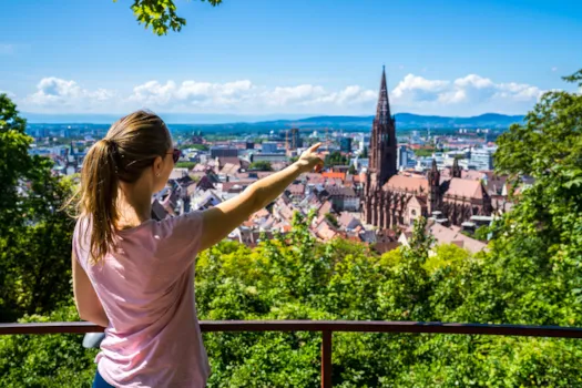 Eine junge Frau, die im Sommer vor blauem Himmel über den Dächern der Altstadt von Freiburg im Breisgau und dem beliebten Münster steht, Baden-Württemberg, Deutschland