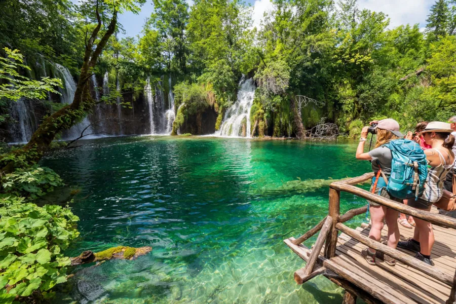 Touristen auf einer Aussichtsplattform, im Hintergrund wunderschöne Wasserfälle – Nationalpark Plitvicer Seen, Kroatien