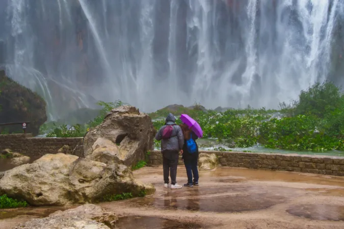 Ein Paar, das unter einem Regenschirm einen Wasserfall in Plitvice bewundert, Nationalpark Plitvicer Seen, Kroatien