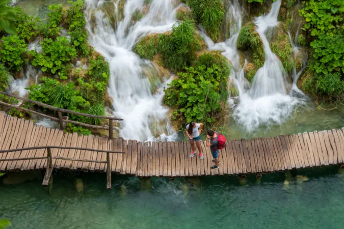 Touristenpaar spaziert auf dem Holzsteg entlang der Wasserfälle, die in den azurblauen Kalu-Erovac-See im Nationalpark Plitvicer Seen in Kroatien hinabstürzen